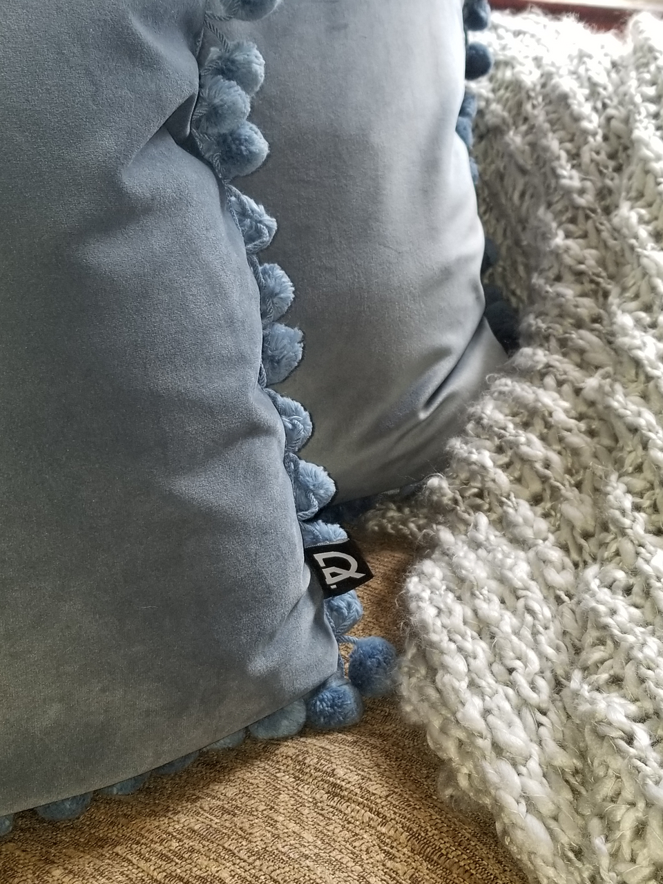 blue velour pillow covers with matching pom pom trim on khaki couch with gray throw cover closeup